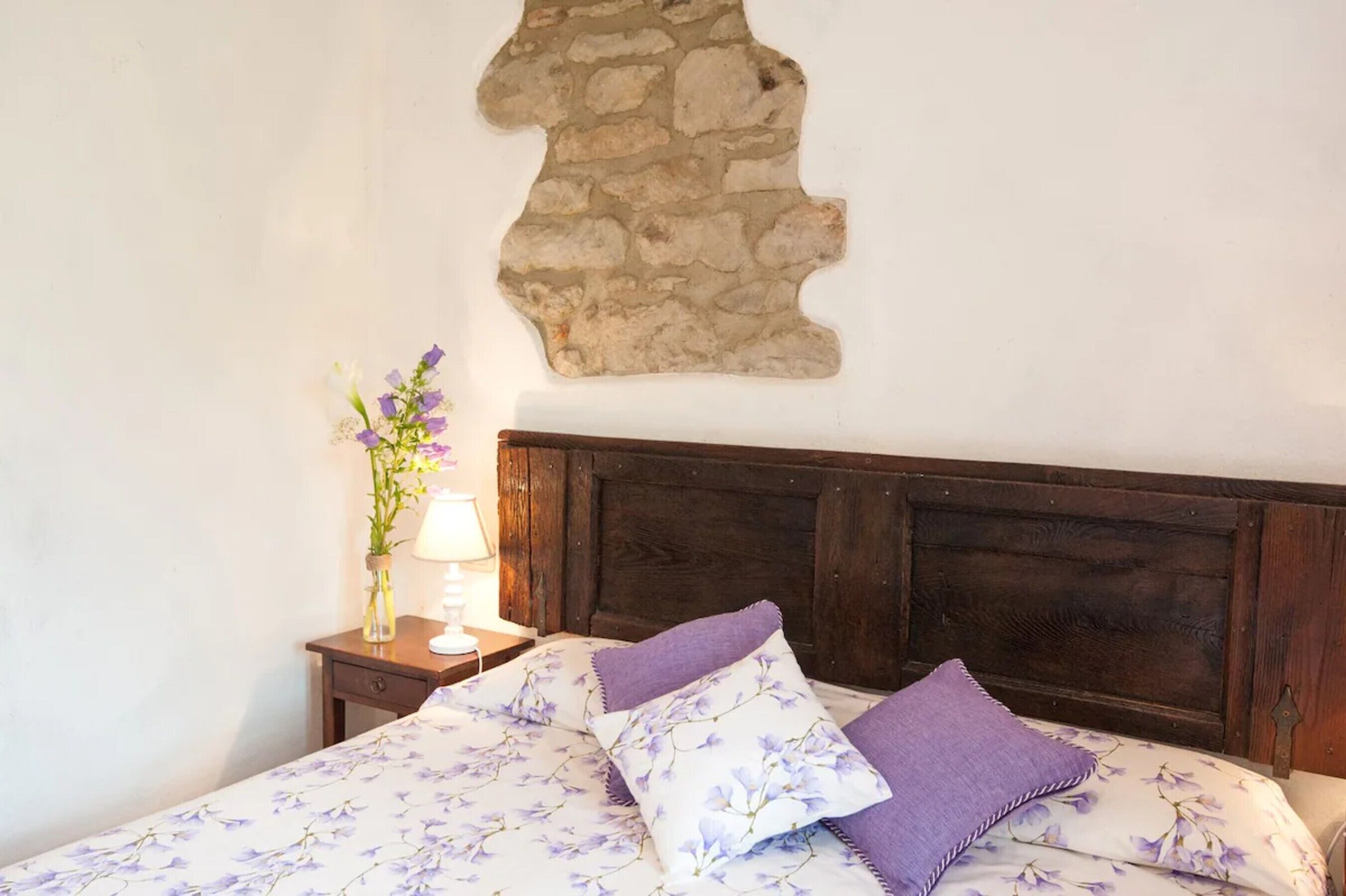 A bed with a floral bedspread, a stone wall, and a nightstand with a lamp and vase of flowers.