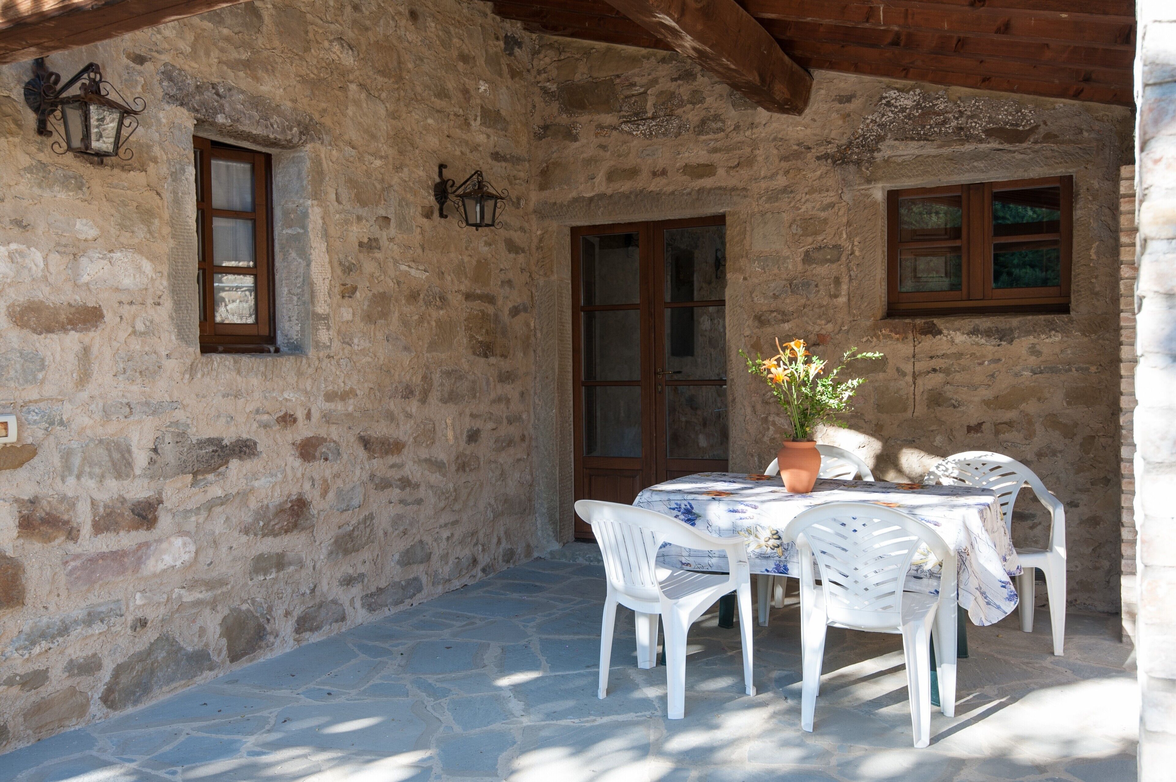 A stone building with a wooden beam ceiling, a table with a floral tablecloth, white plastic chairs, and a potted plant.