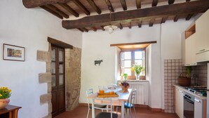 A kitchen with a stone wall, wooden beams, a window with plants, and a dining area with chairs and a table. - Farmhouse "Pilari Appartamento Stalla" with Shared Pool, Private Terrace & Wi-Fi (Cortona)