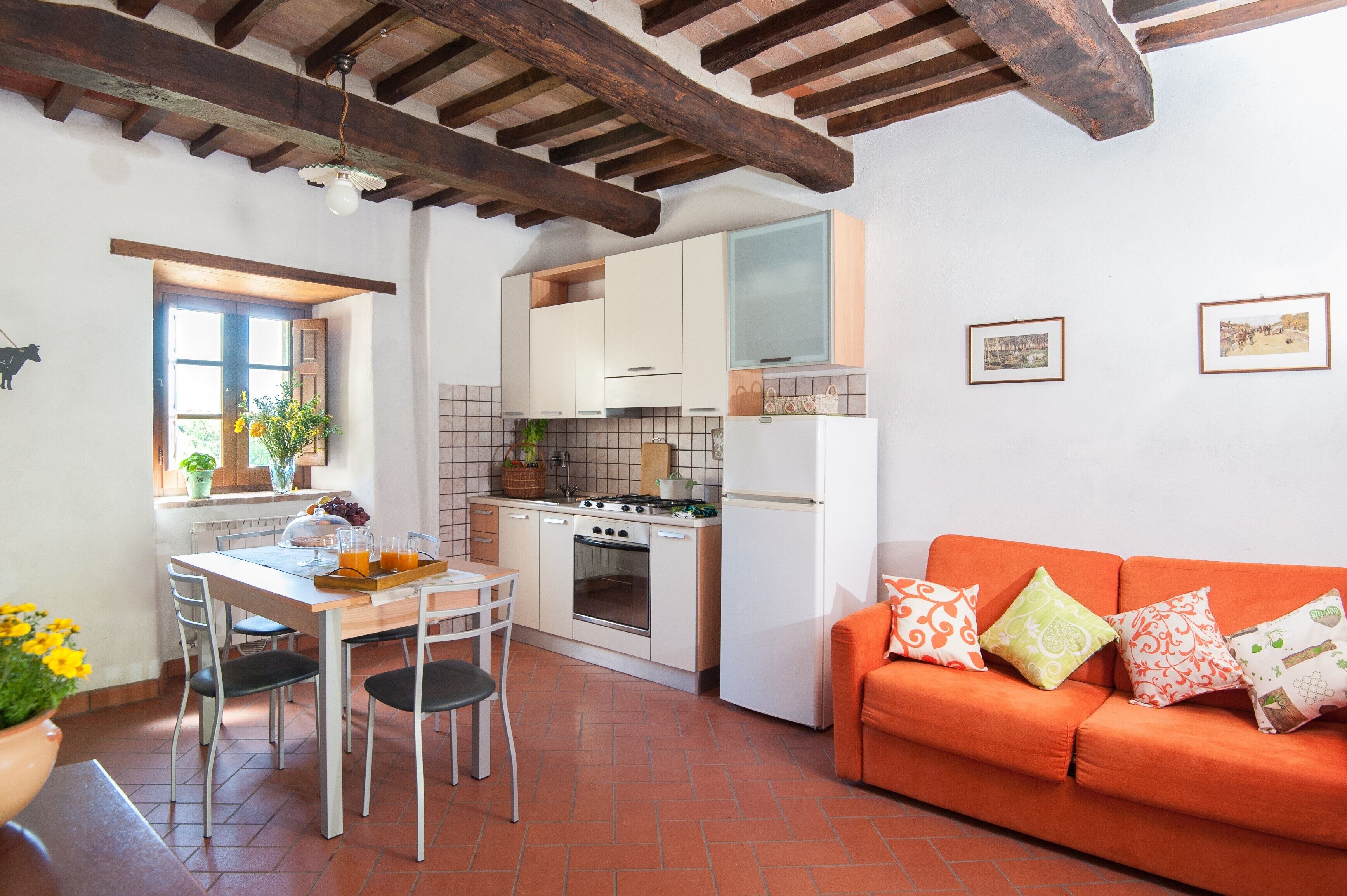 Een keuken met witte kastjes, een fornuis en een koelkast. Er staat een eettafel met stoelen bij een raam met planten. Een oranje bank staat tegen de muur.