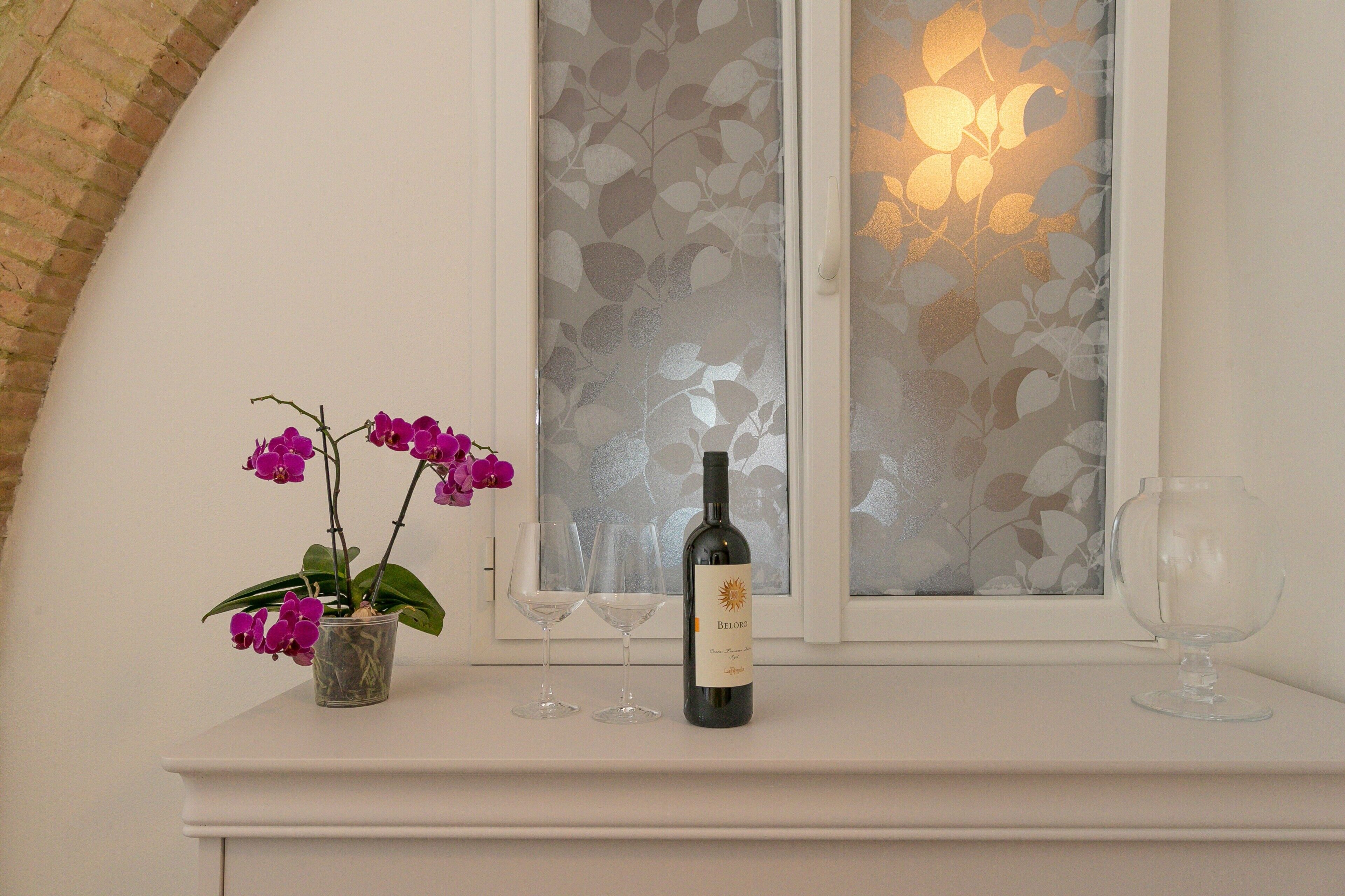 Une bouteille de vin Belardo, deux verres à vin et une orchidée en pot, le tout posé sur une table blanche près d’une fenêtre à vitre dépolie.