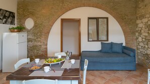 Un coin repas avec une table dressée pour deux personnes, un canapé bleu, un réfrigérateur blanc et un mur en pierre agrémenté d’une porte en arc de cercle.