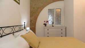 Une chambre à coucher comprenant un lit, une table de chevet, une commode, une fenêtre à vitres dépolies et une arche en briques.