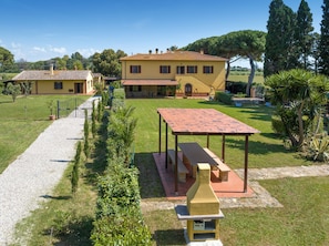 A yellow villa with a tiled roof, a gravel driveway, and a barbecue area.