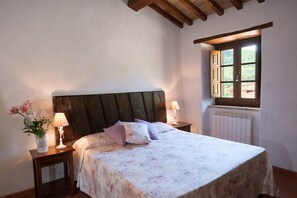 A bedroom with a wooden headboard, a floral bedspread, two bedside lamps, a nightstand with a vase of flowers, and a window with a view of greenery. - Apartment "Pilari Castagnolo" with Mountain View, Shared Pool & Wi-Fi (Cortona)