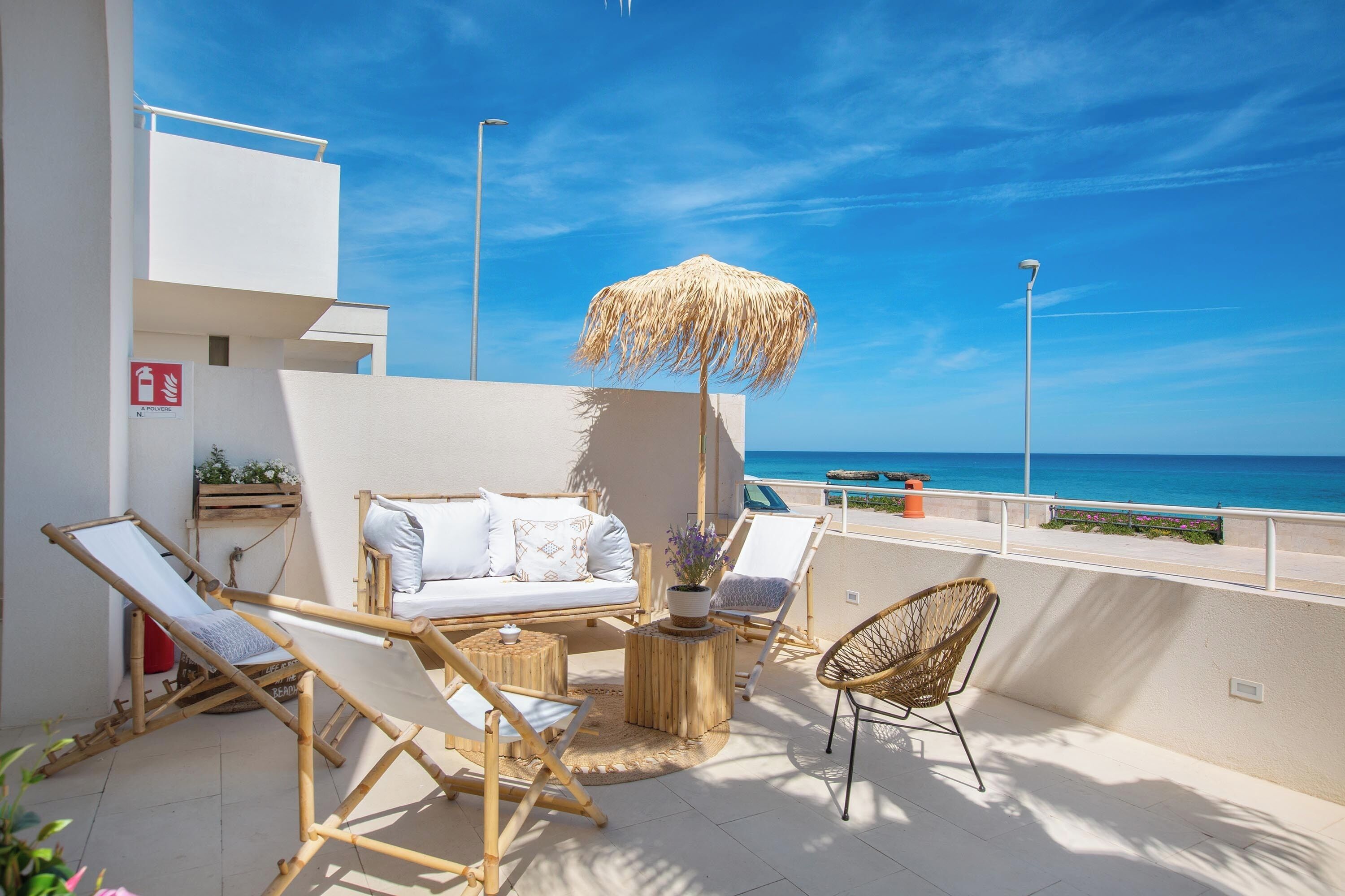 Un balcon avec un canapé blanc, deux fauteuils, une chaise en osier, une petite table ronde ornée d’un pot de plantes et un parasol en chaume.