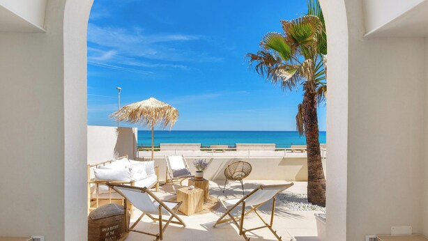 A sunlit outdoor seating area with lounge chairs, a thatched umbrella, and a view of the sea.