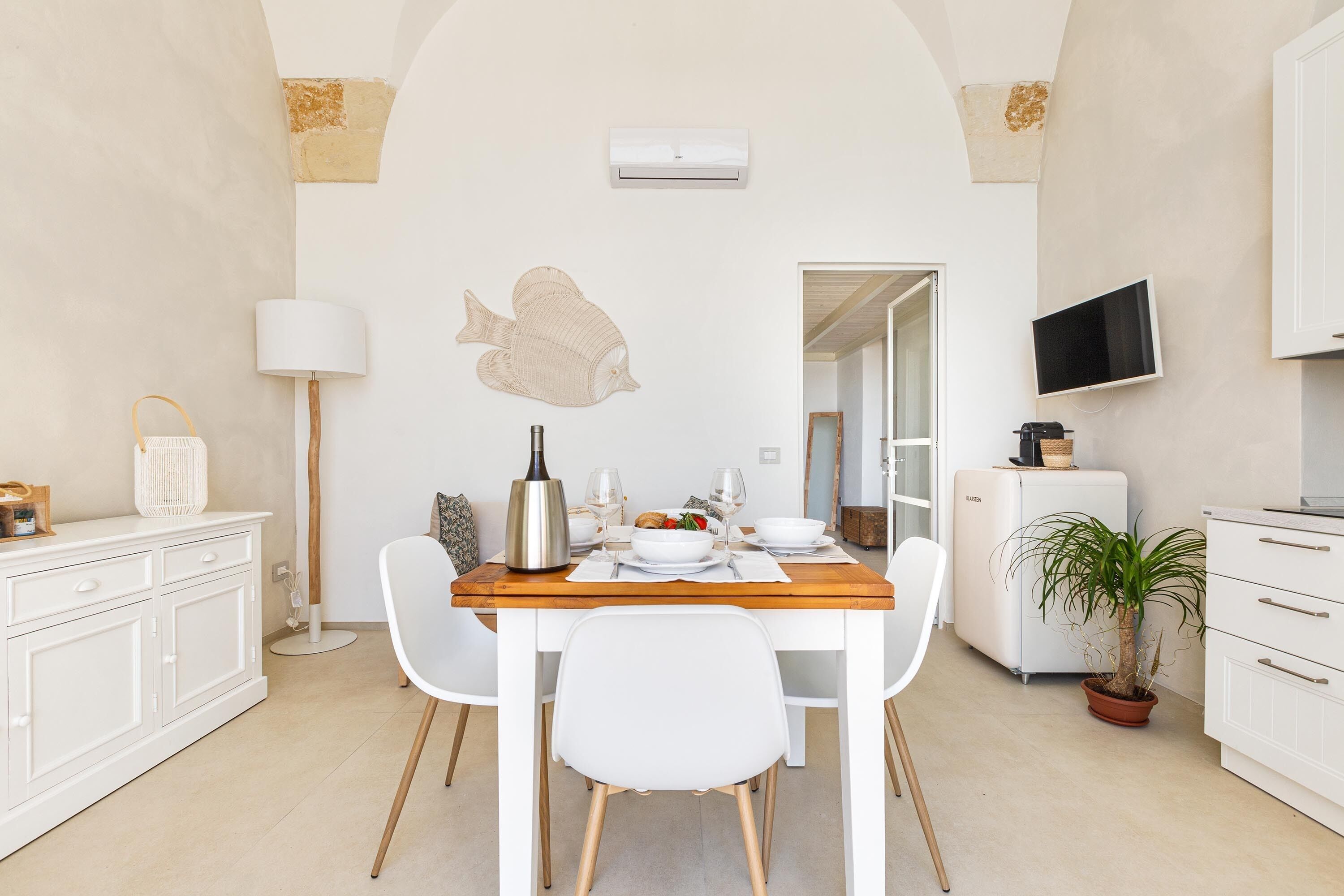 A dining area with a wooden table set for two, white chairs, a white cabinet, a floor lamp, a wall-mounted TV, and a potted plant.