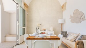 A dining area with a wooden table set for two, white chairs, and a sofa with patterned cushions.