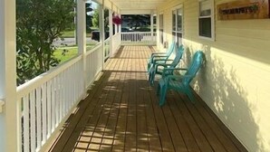 A covered porch with a wooden floor and white railings, featuring two blue chairs and a small table. - The Yellow House-Three bedroom within walking distance of Lake Michigan. (Ludington)