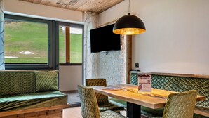 Ein Zimmer mit Holzboden, einem großen Fenster, einer grünen gepolsterten Bank, einem Holztisch und einer schwarzen Pendelleuchte.