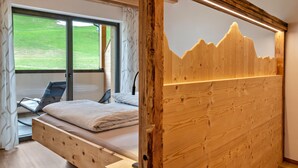 Ein Schlafzimmer mit Holz-Kopfteil, einem Bett mit weißer Matratze und Kissen, einem Sessel und einem großen Fenster mit Blick auf eine grüne Landschaft.