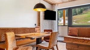 Una habitación con una mesa de comedor de madera, sillas acolchadas, un sofá, un televisor de pantalla plana y un ventanal con vistas a un paisaje verde.