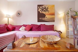 A living room with a sofa, a coffee table, a painting, a lamp, and a bookshelf. - Holiday Home 'Oasis Tamarindo Sport & Relax' with Shared Pool, Private Terrace & Wi-Fi (Corralejo)