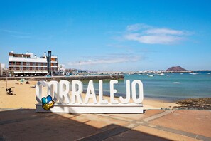 A beachside sign with 'CORRALEJO' and a cartoon character, a building with balconies, and boats in the water. - Holiday Home 'Oasis Tamarindo Sport & Relax' with Shared Pool, Private Terrace & Wi-Fi (Corralejo)