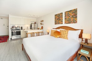 A hotel room with a bed, a wooden headboard, a nightstand, a lamp, a bedside table, a kitchen area with a stove, refrigerator, and shelves.
