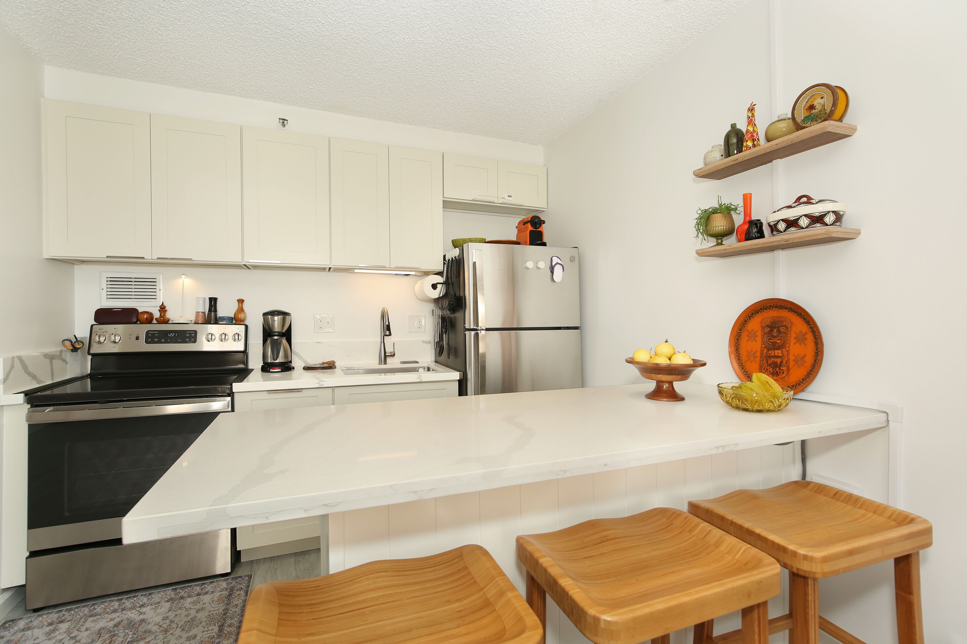 Cocina moderna con armarios blancos, electrodomésticos de acero inoxidable, una isla central con taburetes y objetos decorativos en estantes.