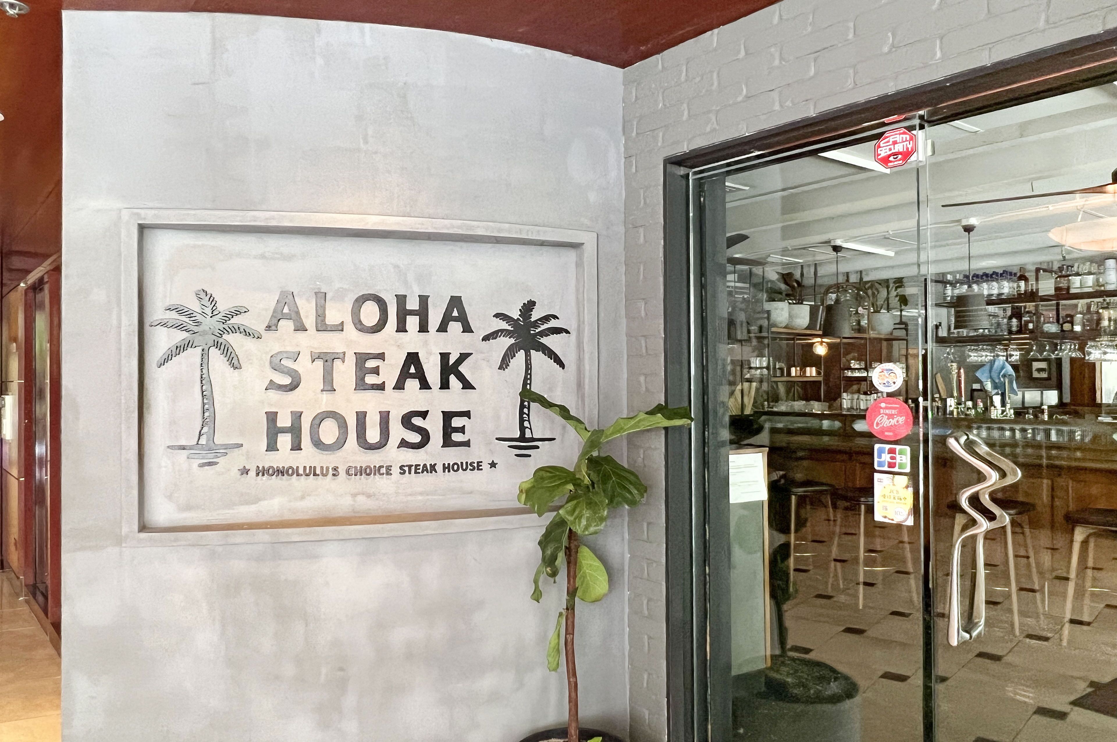 Entrada de un restaurante con un letrero que dice 'ALOHA STEAK HOUSE' y un gráfico de una palmera.