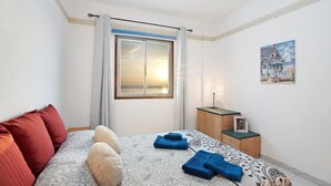 A bedroom with a bed, pillows, a nightstand, a lamp, a window, and a painting on the wall.