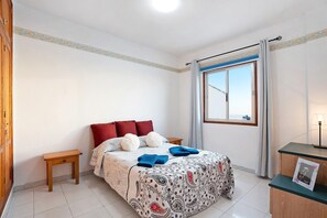 Une chambre à coucher avec un lit, une petite table de chevet en bois, un bureau avec une lampe et une fenêtre avec des rideaux.