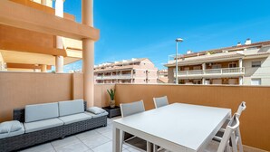 Une terrasse extérieure avec une table blanche et un canapé gris, entourée de murs de couleur beige et sous un ciel d’un bleu limpide.