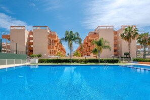 En swimmingpool med en klar, blå vandoverflade, omgivet af moderne lejlighedskomplekser med altaner og palmer.