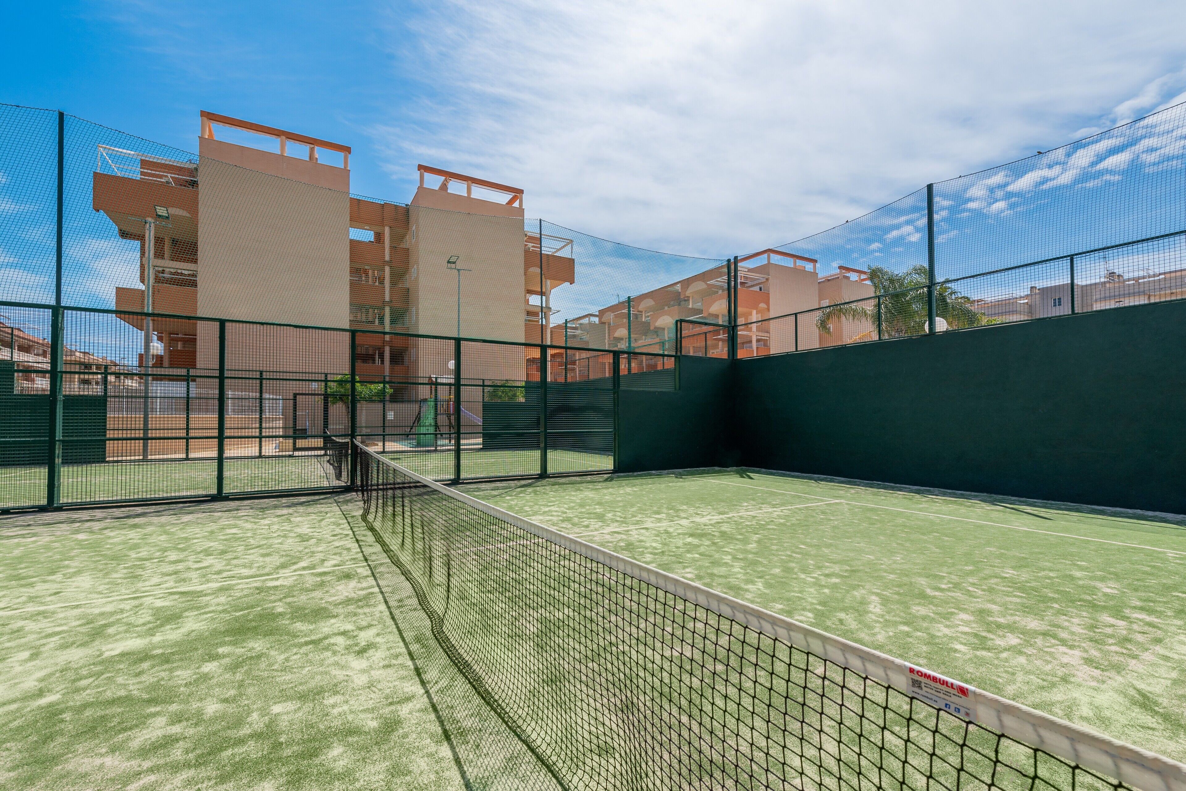 Un court de tennis intérieur, doté d’un filet, entouré d’une clôture et bordé de bâtiments adjacents.