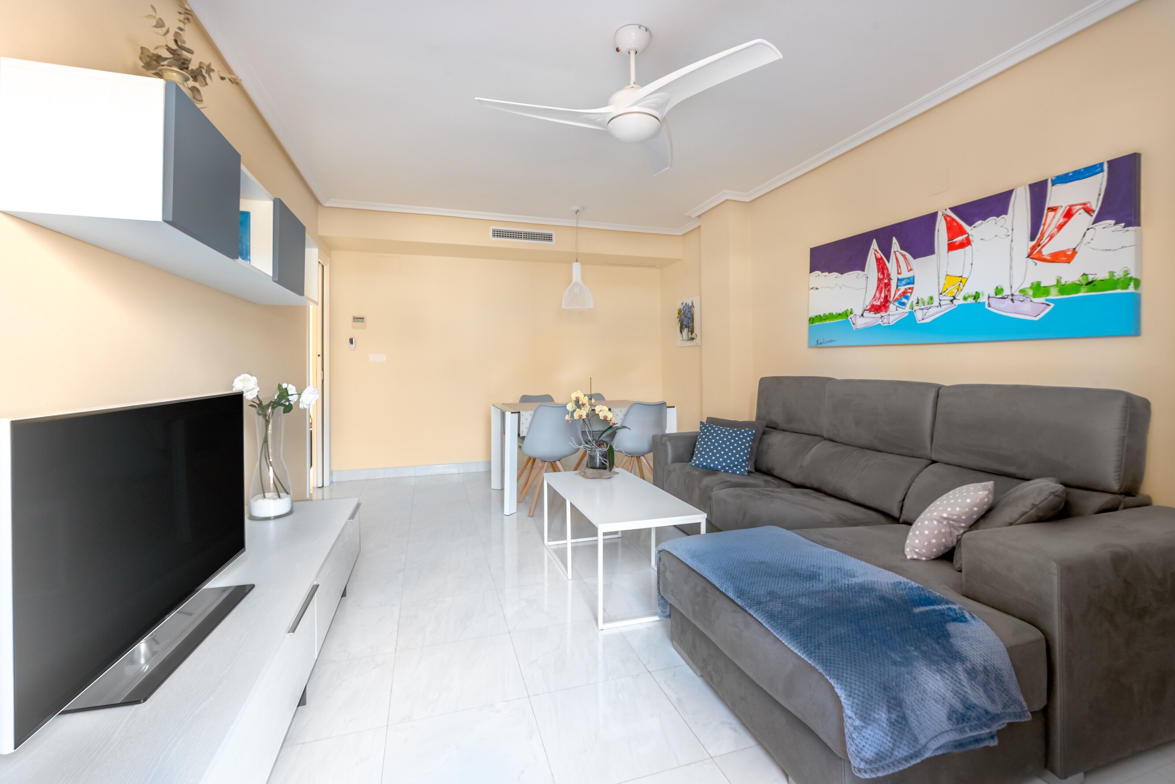 Un salon moderne avec un téléviseur à écran plat, un canapé gris, une table basse blanche, un ventilateur de plafond et un tableau représentant un voilier accroché au mur.
