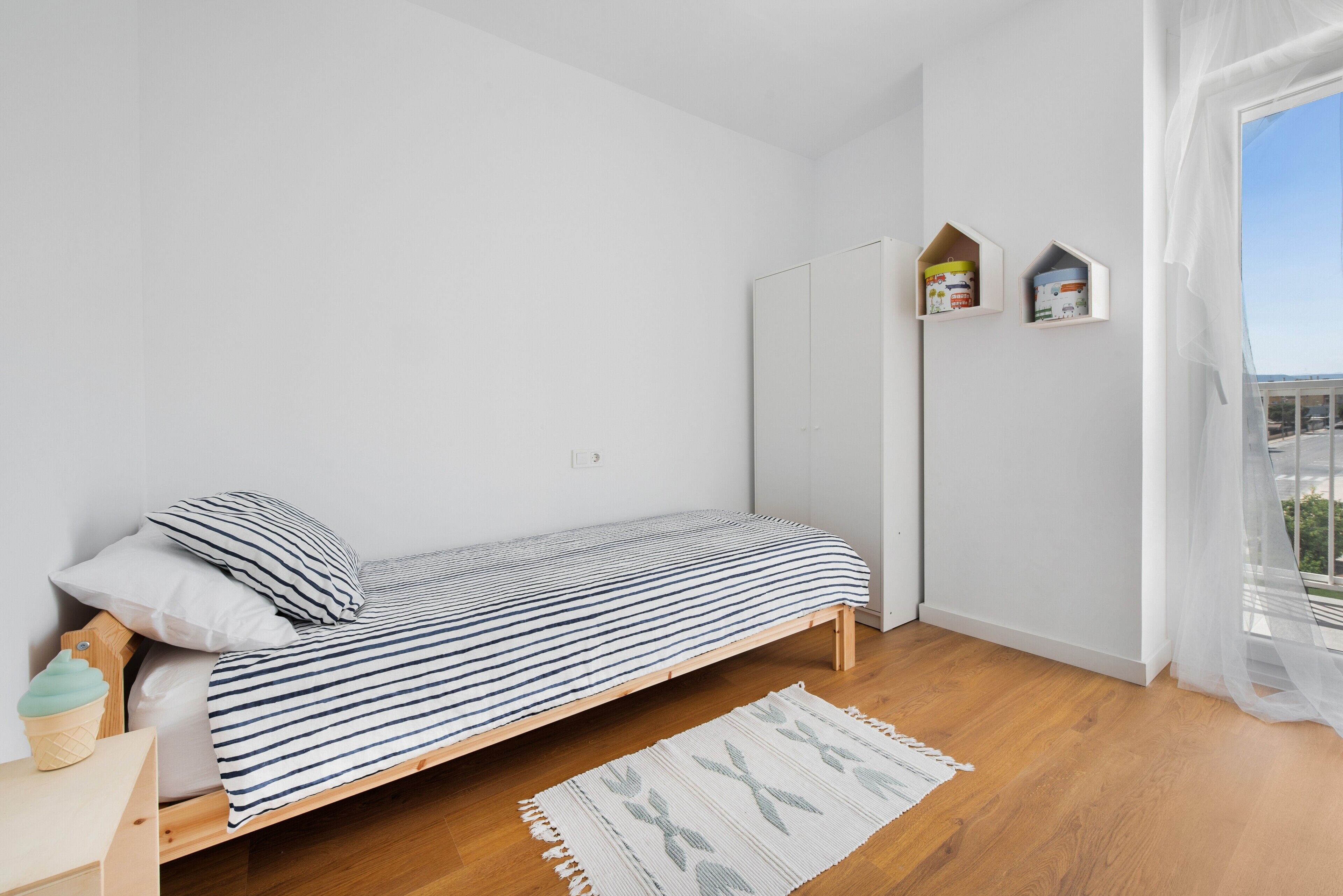Una cama individual con colcha a rayas, una mesita de noche con lámpara, un pequeño estante con libros y vistas al exterior a través de una ventana.