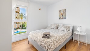 Une chambre avec un lit, une fenêtre donnant sur une piscine et des bâtiments, un tableau accroché au mur et une table de chevet avec une lampe.