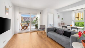 Un salon moderne avec un canapé gris, un téléviseur à écran plat, un coin repas comprenant une table et des chaises, et un balcon offrant une vue sur les immeubles.
