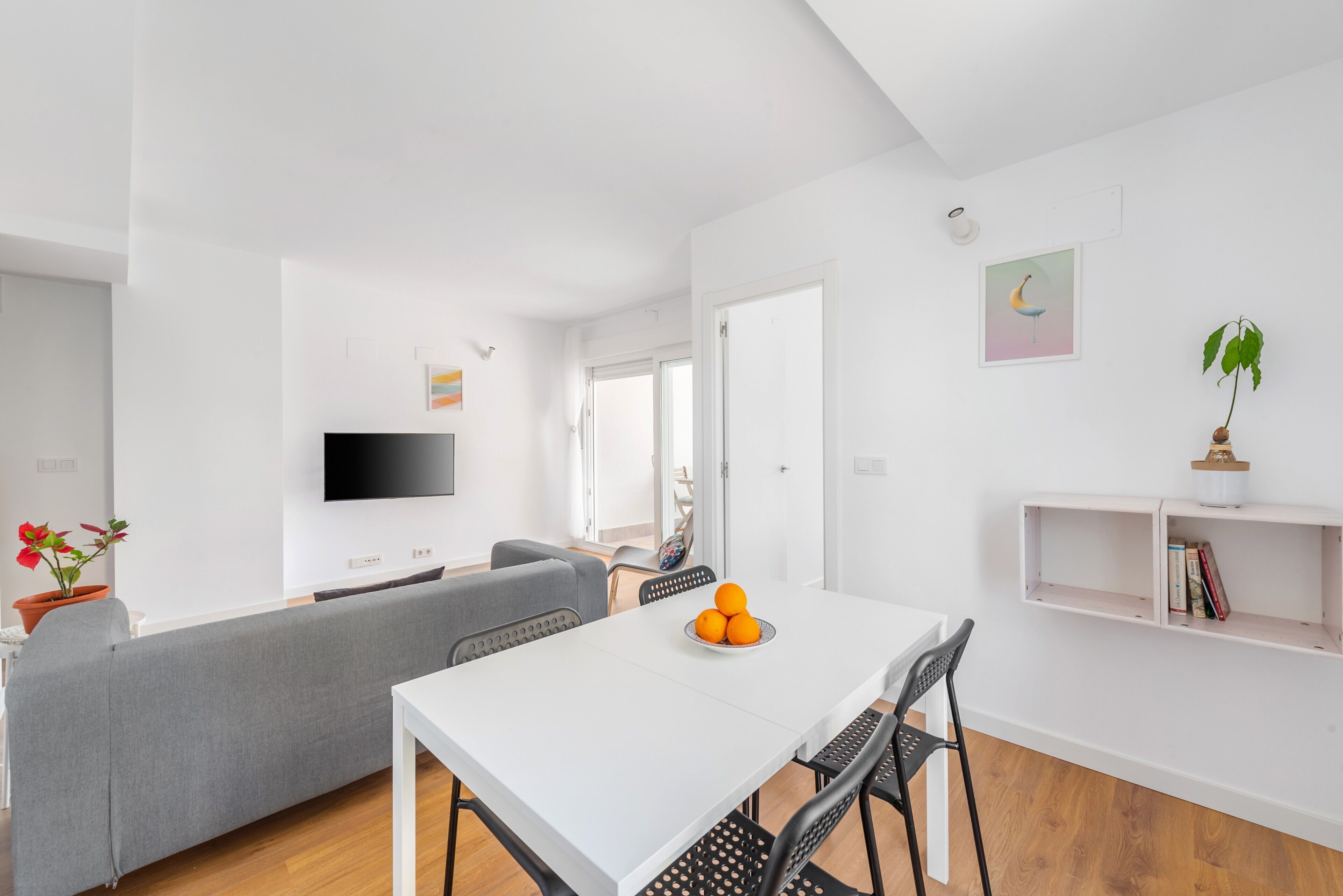 A modern living room with a white sofa, a dining table with chairs, a flat-screen TV, and a small plant on a shelf.