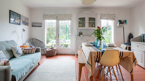 Un salón luminoso con un sofá, una mesa de comedor con sillas y una zona de cocina con armarios y una ventana.