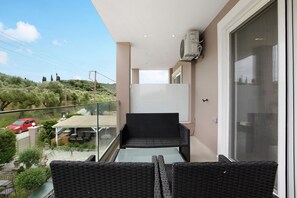 Un balcon agrémenté de meubles en osier, d’une table en verre et offrant une vue sur une voiture rouge et de la verdure.