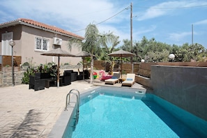 Une piscine avec des chaises longues et des parasols, une maison avec un toit en tuiles et un mur en pierre.