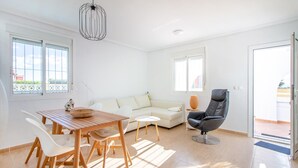 A bright, modern living room with a wooden dining table, white chairs, a beige sofa, and a black leather chair.