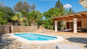 Une piscine aux bords recouverts de pierres, un patio abrité avec des sièges et une végétation luxuriante tout autour.