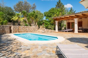 Una piscina con baldosas de piedra, un porche cubierto con asientos y vegetación circundante.