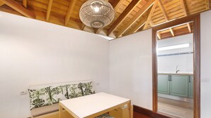 A room with a wooden ceiling, a white table, a bench with a leaf-patterned cushion, and a view of a kitchen through a doorway.