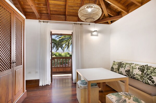 A room with a wooden ceiling, a white table, a sofa with leaf-pattern cushions, and a view of palm trees through a doorway.