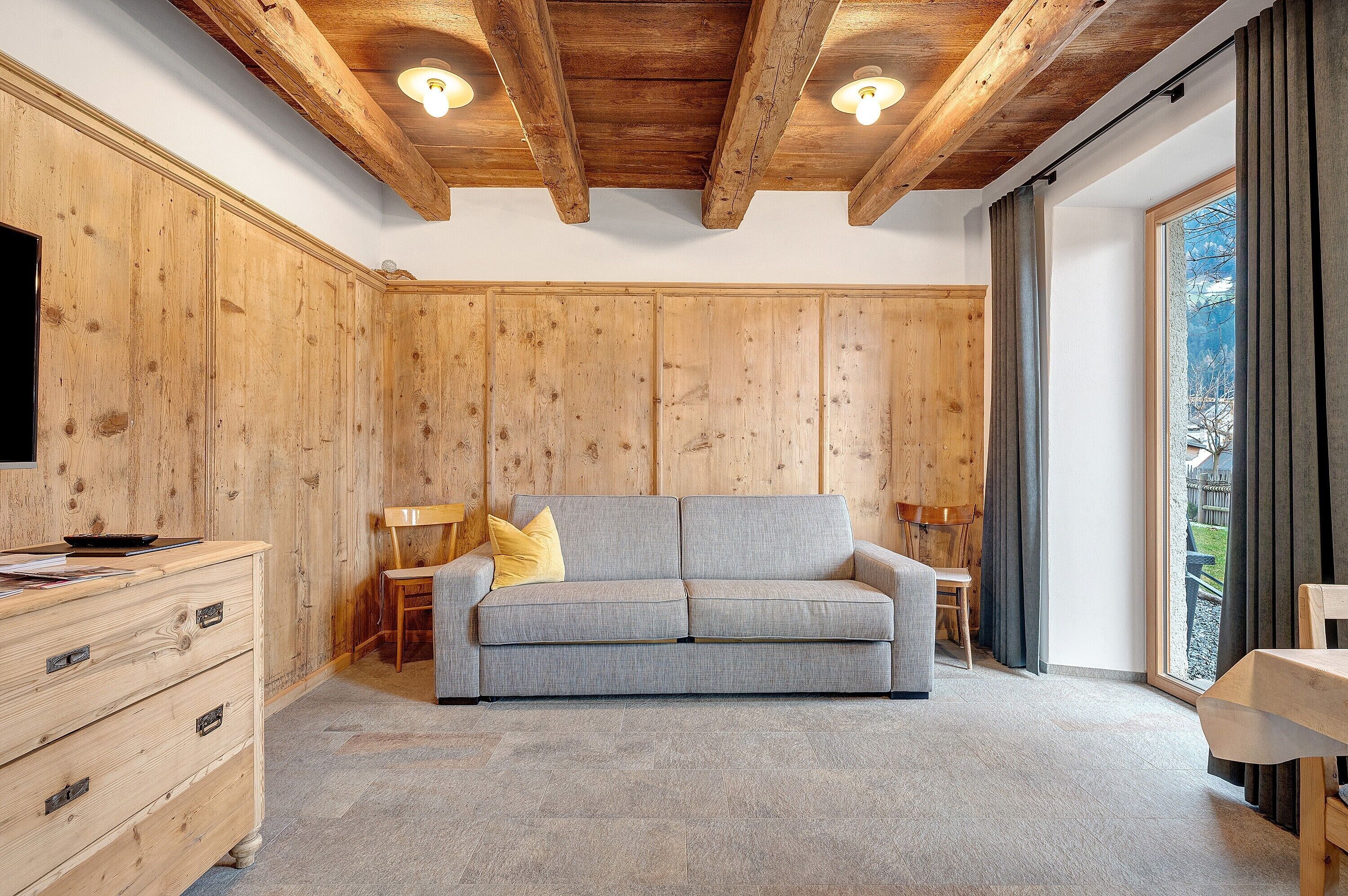 Une pièce avec un plafond en bois, un canapé gris, une commode en bois et une grande fenêtre avec des rideaux.