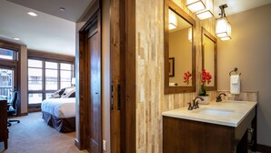 A hotel room with a large bed, a wooden wardrobe, a bathroom with a double sink and a large mirror, and a view of the outside through large windows. - Luxury 2BD Village at Northstar Residence w/ Ski Valet (Truckee)