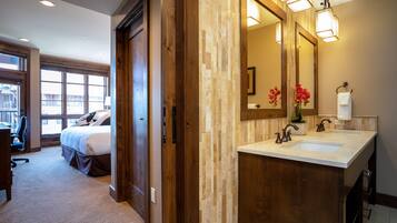 Une chambre d’hôtel avec un grand lit, une armoire en bois, une salle de bain dotée d’un lavabo double et d’un grand miroir, et une vue sur l’extérieur grâce à de grandes fenêtres.