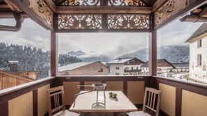 Een houten balkon met een tafel en stoelen, uitkijkend op een besneeuwd dorp en bergen.