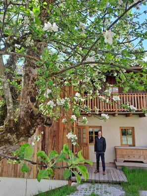 Una persona parada frente a una casa de madera con balcón, rodeada de árboles en flor.