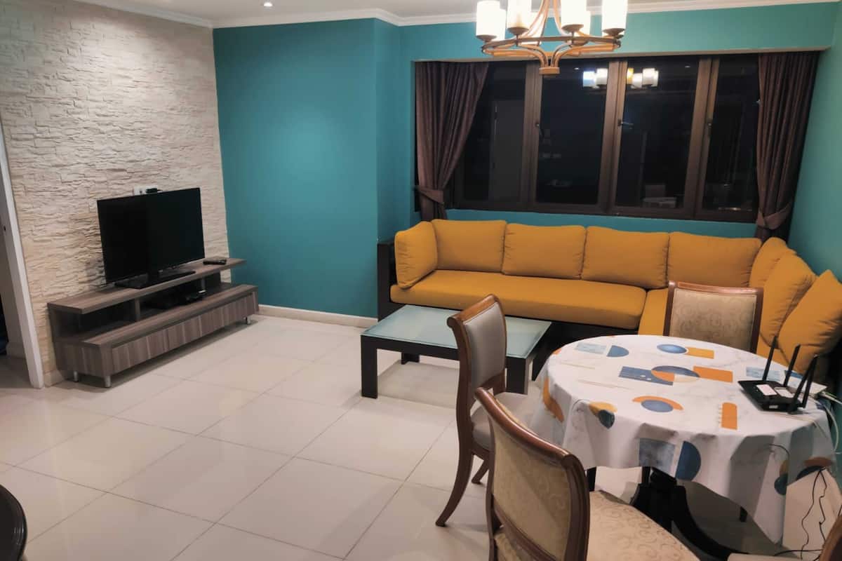 A living room with a teal wall, a sofa, a TV on a wooden stand, and a dining table with chairs.