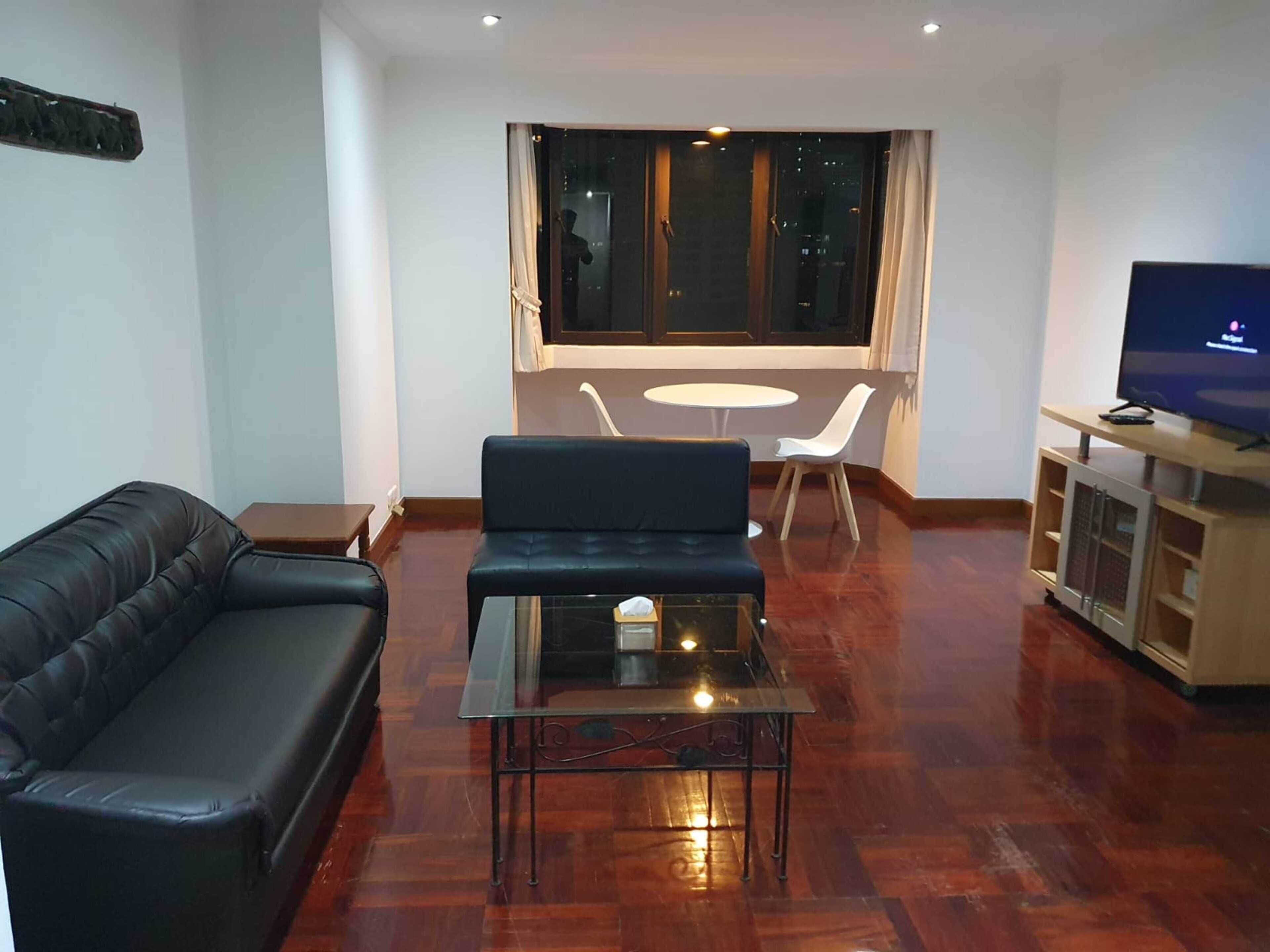 A living room with a black leather sofa, a glass coffee table, a TV on a wooden stand, and a view of the city through large windows.