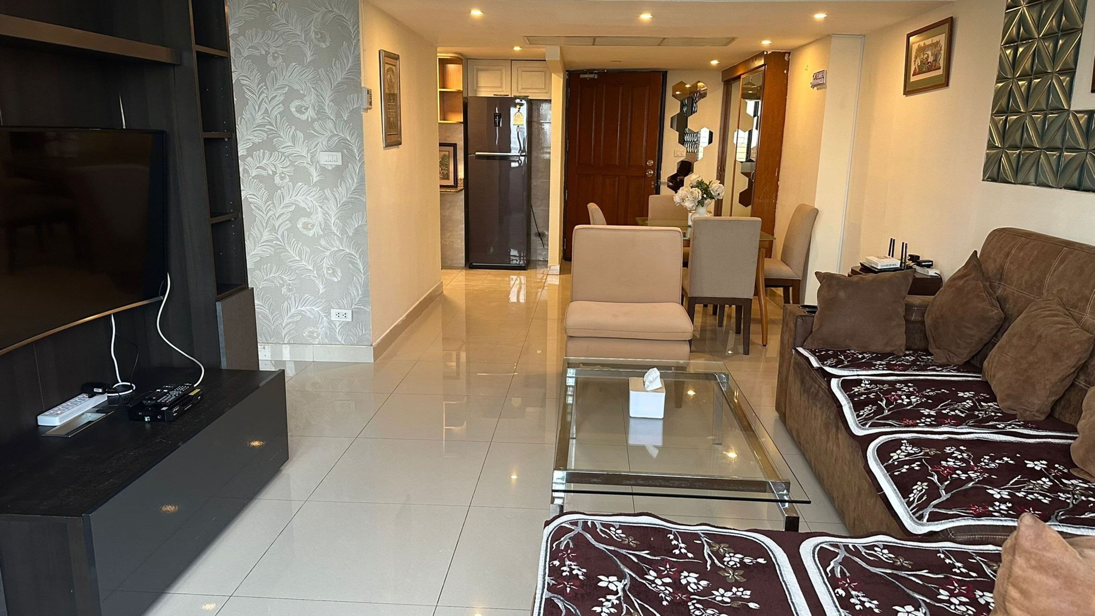 A modern living room with a flat-screen TV, a glass coffee table, a sofa, and a dining area with chairs and a table.