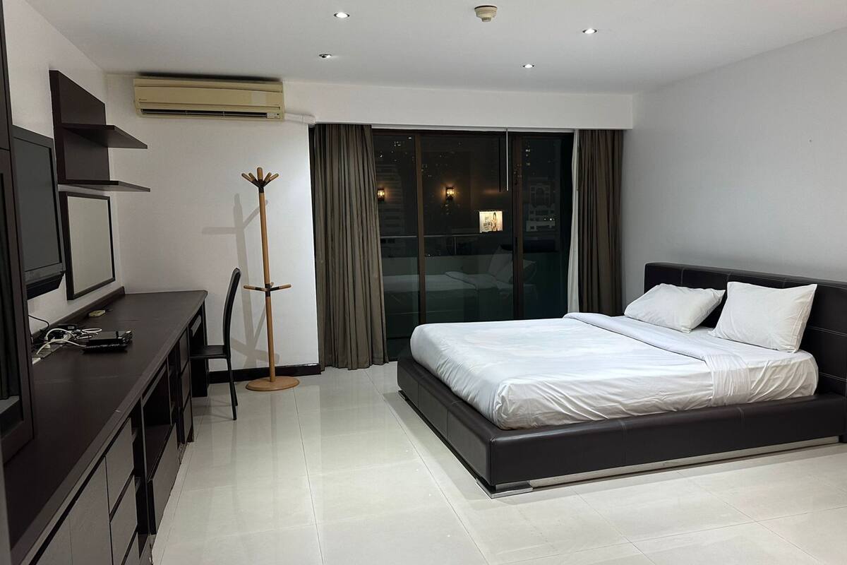 A modern hotel room with a large bed, a desk with a chair, a standing lamp, and a television.