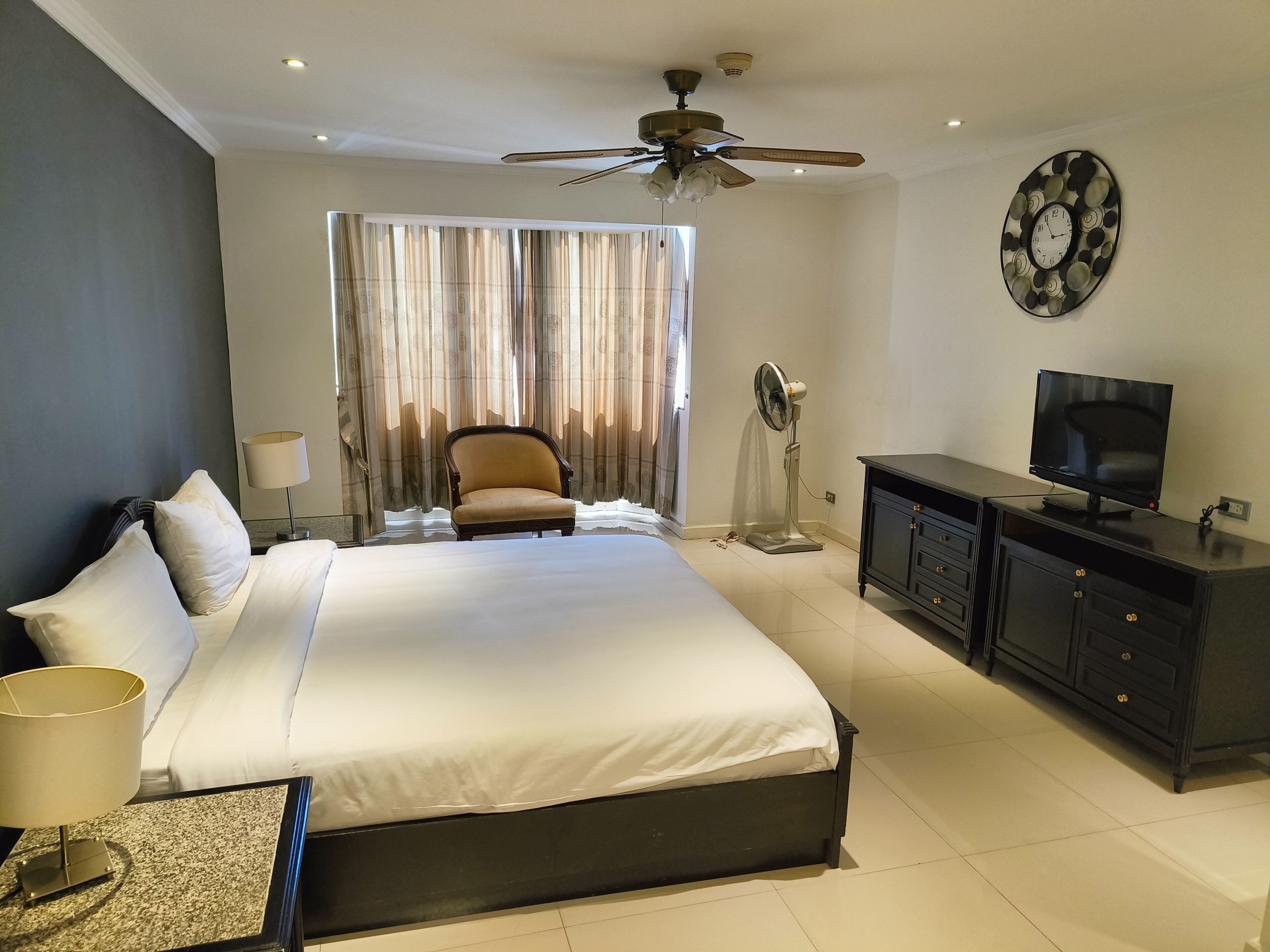 Habitación de hotel con una cama grande, ventilador de techo, televisor, escritorio con lámpara y una silla.